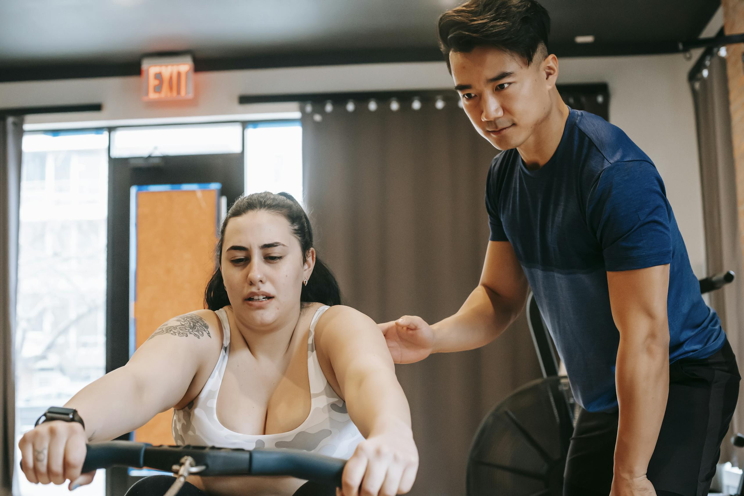 Young handsome ethnic experienced trainer supporting plus size woman working out on rowing machine in gym
